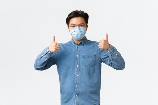 Covid-19, preventing virus, and social distancing at workplace concept. Confident asian man in shirt and medical mask showing thumbs-up, ensuring safety of employees during coronavirus.