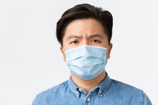 Coronavirus, social distancing and lifestyle concept. Close-up of gloomy disappointed asian man in medical mask looking with condemn, frownign displeased, standing white background.