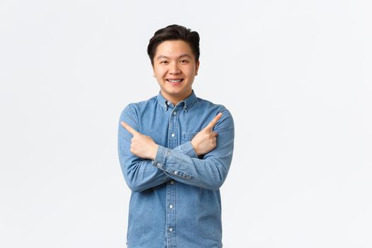 Smiling cheerful asian man with braces making announcement. Guy pointing fingers sideways at left and right variants, showing few options, recommend stores, standing white background.