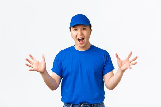 Portrait of excited and euphoric asian delivery man in blue uniform react to wonderful news, shaking hands happy and thrilled, telling big announcement, rejoicing over white background.