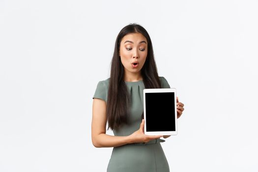 Small business owners, startup and work from home concept. Amazed and impressed asian woman in dress looking at digital tablet screen with astounded expression, checking out new items in stock.