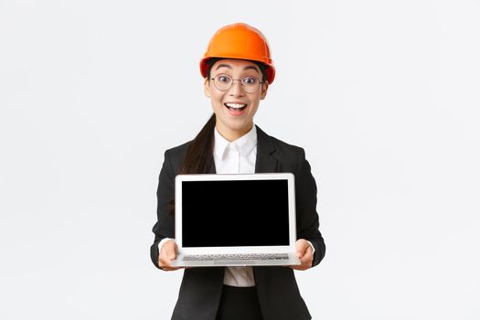 Excited happy asian female engineer, industrial manager in business suit and safety helmet showing laptop screen to investors or clients, amazed with great profit chart, introduce diagram.
