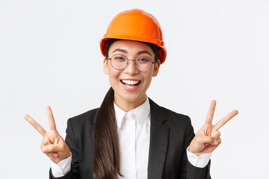 Close-up of cheerful successful female asian engineer, construction architect in safety helmet and business suit showing peace signs and smiling kawaii, standing white background.