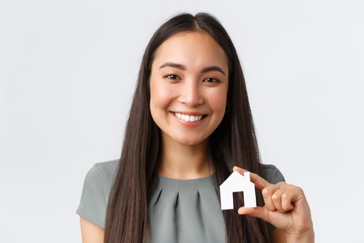 Insurance, loan, real estate and family concept. Close-up of happy smiling asian woman showing small paper house miniature in hands and looking at camera, found perfect apartment, white background.