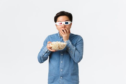Leisure, lifestyle and people concept. Astonished asian guy in 3d glasses munching popcorn excited looking at TV, watching movie on wide screen, attend cinema, standing white background.