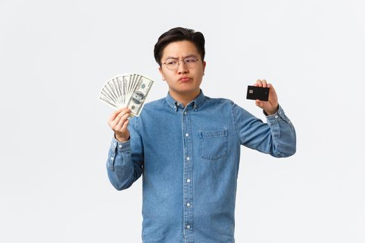 Perplexed asian businessman in glasses holding cash and money, looking doubtful at money, thinking use contactless payment during coronavirus pandemic, standing white background.