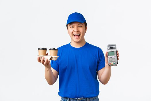 Contactless shopping, payment and delivery concept. Cheerful smiling male courier in blue uniform holding takeaway coffee and POS terminal for client to pay for their order, white background.