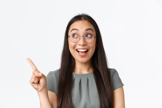 Close-up of upbeat and excited asian female in glasses, looking and pointing finger upper left corner with fascinated cheerful face, checking-out awesome promotion, standing white background.