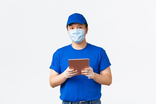 Online shopping, covid-19, preventing virus and delivery concept. Young smiling asian courier in medical mask and gloves checking address or client info on digital tablet app, white background.