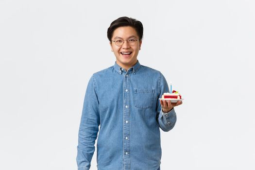 Celebration, holidays and lifestyle concept. Smiling handsome asian man with braces looking upbeat, holding piece of birthday cake with candle, celebrating b-day, standing white background.