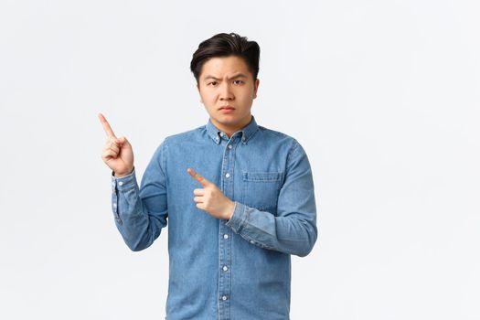 Skeptical and angry asian man pointing fingers upper left corner, showing bad news, frowning disappointed, scolding for something, standing white background displeased, making statement.