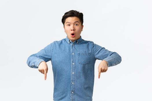 Shocked and impressed asian guy in blue shirt, pointing fingers down and looking at camera speechless, asking question about product, found something interesting, standing white background.