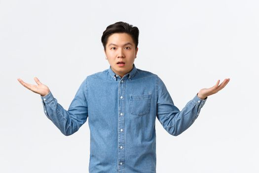 Confused and puzzled asian man in shirt cant understand what happening, raising hands sideways and shrugging, waiting explanation, being wondered, standing questioned white background.