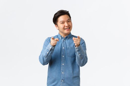 Confident and cheerful asian young man with braces, smiling, encourage person, pointing fingers at camera as choosing someone or congratulating lucky winner, standing white background.