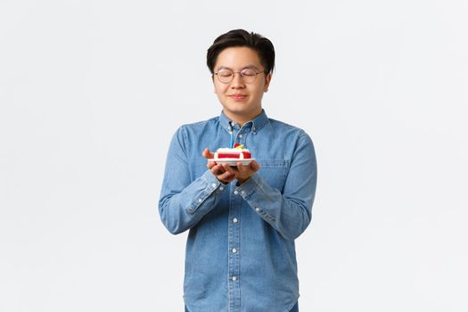 Celebration, holidays and lifestyle concept. Dreamy cute asian man in glasses close eyes as making wish and blowing lit candle on birthday cake, celebrating b-day, standing white background.