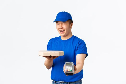 Contactless shopping, payment and delivery concept. Cheerful friendly, smiling asian courier in blue uniform bring order to client home, handing pizza box and POS terminal, white background.