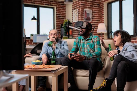 Multiethnic group of friends using virtual reality glasses to play video games on television. Having fun with beer bottles and snacks at house oarty, playing competition with vr goggles.