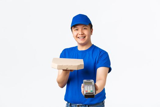 Contactless shopping, payment and delivery concept. Cheerful friendly, smiling asian courier in blue uniform bring order to client home, handing pizza box and POS terminal, white background.