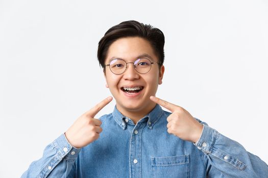 Orthodontics and stomatology concept. Close-up of satisfied asian guy, dental clinic client smiling happy and pointing at his dental braces, standing white background, recommend quality.