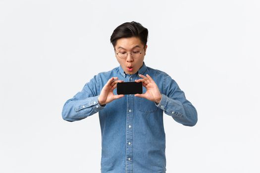 Impressed asian guy react to new movie trailer, showing friends mobile phone screen, stare impressed at smartphone screen, astonished with graphics of game, standing white background.