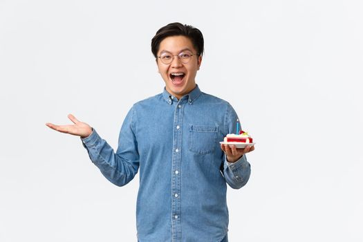 Celebration, holidays and lifestyle concept. Excited and amused happy asian guy looking with rejoice, smiling and holding birthday cake, attend b-day party, standing white background.