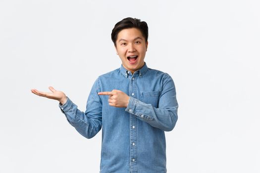 Smiling cheerful cute asian man making announcement, demonstrate new product on hand, pointing finger at blank space for your advertisement, standing amused over white background.