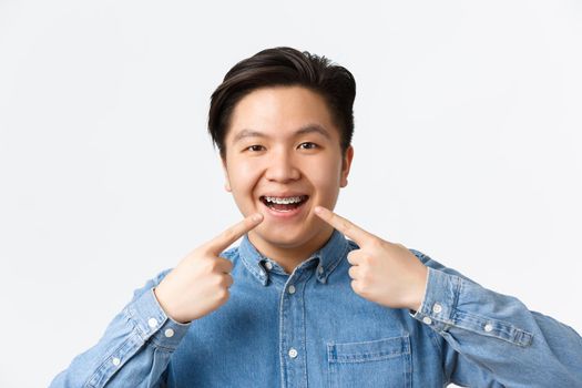 Orthodontics and stomatology concept. Close-up of happy smiling asian man pointing fingers at dental braces on teeth with pleased expression, recommend dentist clinic, standing white background.
