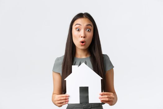 Insurance, loan, real estate and family concept. Surprised asian woman in dress, holding paper house in hands react to big announcement, found excellent apartment for cheap price, white background.