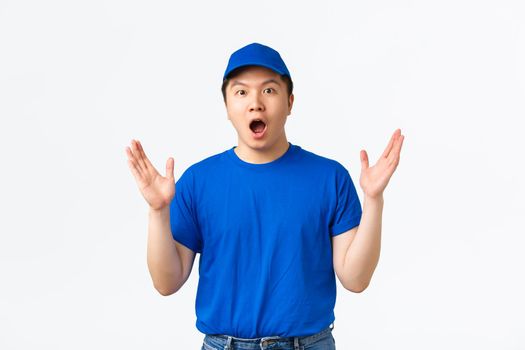 Surprised and excited courier in blue uniform react to awesome news, big announcement in mail company. Impressed delivery guy shaking raised hands and gasping amazed, white background.