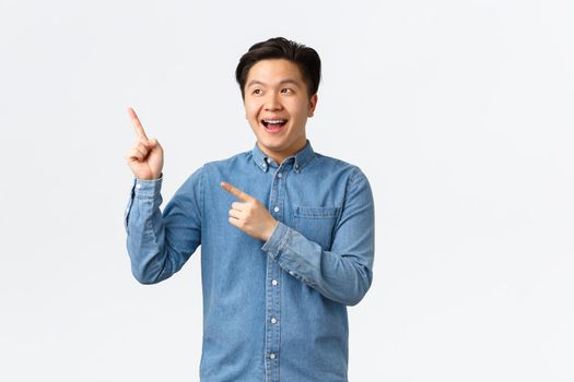 Outgoing cheerful asian man looking and pointing fingers upper left corner with excited smile, showing big news, making announcement, promote link or online store, white background.