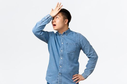 Annoyed and bothered asian male freelancer forget something, rolling eyes irritated and punch forehead, making facepalm gesture as hear lame offer, standing white background displeased.