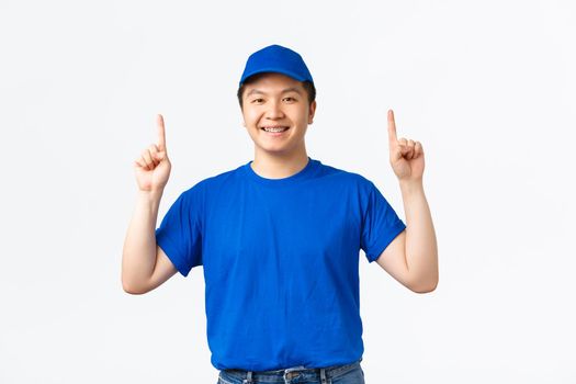 Upbeat smiling asian courier with braces, wearing blue company uniform, pointing fingers up, inform clients about discounts on shipping, special offers, standing white background.