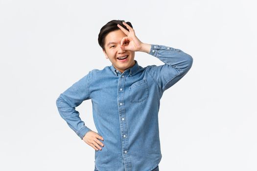 Smiling cheerful and satisfied asian man showing okay gesture over eye, looking through with pleased expression, assure everything good, recommend excellent service, guarantee.
