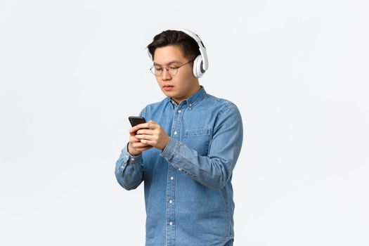 Lifestyle, leisure and technology concept. Serious-looking asian teenage guy in glasses, listening music on wireless headphones and messaging friend, using mobile phone adjust earphones sound.