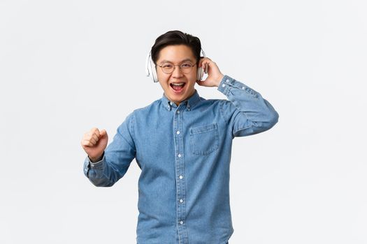 Lifestyle, leisure and technology concept. Joyful happy asian man in glasses, dancing carefree to awesome sound of music, great earphones quality, wearing wireless headphone, enjoying listening song.