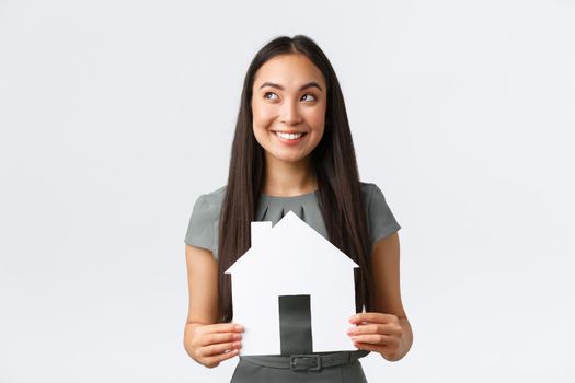 Insurance, loan, real estate and family concept. Thoughtful smiling asian woman in dress, holding paper house in hands and looking away while thinking, imaging perfect home, white background.