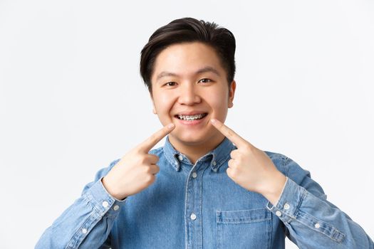Orthodontics and stomatology concept. Close-up shot of happy smiling asian man recommend dental clinic, pointing fingers at teeth braces with satisfied expression, white background.