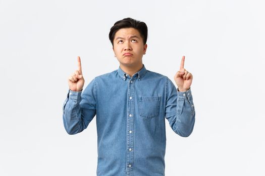 Skeptical and unamused asian man in blue shirt looking and pointing fingers up with disappointed expression, pouting distressed, complaining over something bad, standing white background.