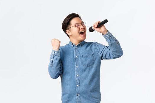 Lifestyle, leisure and people concept. Carefree happy asian man enjoying singing at karaoke, holding microphone and fist pump in delight, performing over white background.