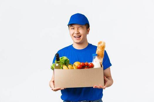 Online shopping, food delivery and shipment concept. Winking cute asian male courier in blue uniform provide fast shipment, bring groceries order to client house, handing box and smiling.