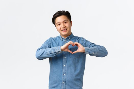 Smiling cute asian man in blue shirt showing heart gesture, express sympathy or care, in love with someone, open own feelings towards person, standing white background. Copy space
