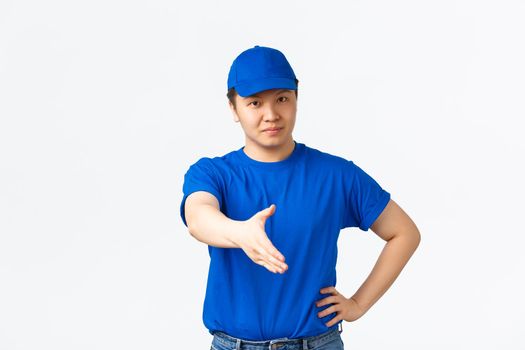 Friendly cheerful asian courier in blue uniform smiling confident, extend hand for handshake, greeting customer, making deal. Delivery man saying hello and standing white background. Copy space