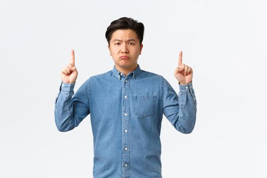 Disappointed gloomy and upset asian man inform about bad sad news, pointing fingers up and looking at camera displeased, complaining, feeling uneasy, standing white background.