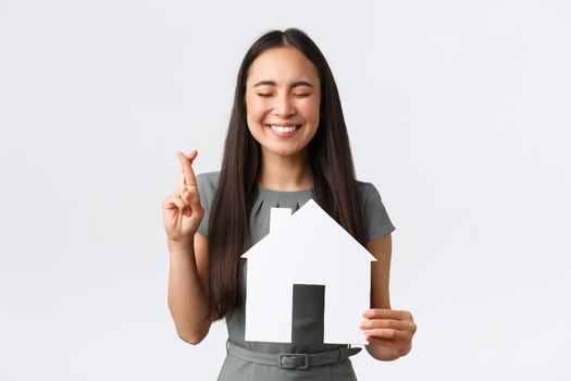 Insurance, loan, real estate and family concept. Hopeful smiling asian woman praying finding new apartment, cross fingers good luck, making wish with closed eyes, holding paper house.