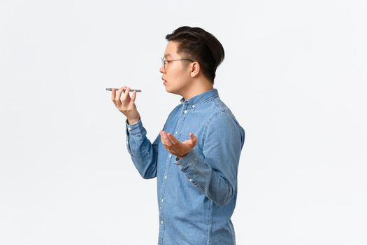 Profile of busy young asian male entrepreneur, freelancer record voice message, talking into phone speaker, making notes on recorder, having conversation, standing white background.