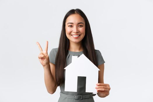 Insurance, loan, real estate and family concept. Smiling attractive asian woman selling or buying apartment, showing peace kawaii sign and paper house, saleswoman showing home, white background.