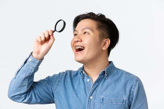 Close-up of amused and excited asian young man with braces, smiling satisfied, found something interesting, looking through magnifying glass at upper left corner, white background.