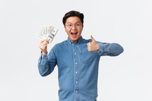 Excited smiling asian man with braces and glasses, showing thumbs-up and waving money, receive paycheck, freelancer got job with good payment, holding cash and looking satisfied.