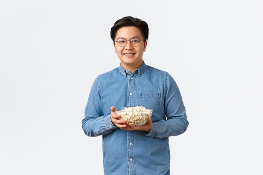 Leisure, lifestyle and people concept. Modest cute asian guy with braces and glasses holding popcorn and smiling at camera, ready for watching premier on tv, standing white background.