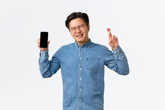 Hopeful asian guy with braces, close eyes and cross fingers good luck, showing smartphone screen, waiting for reply on dating app, begging god, standing white background and making wish.